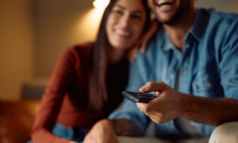Couple laughing while watching TV, man holds remote.