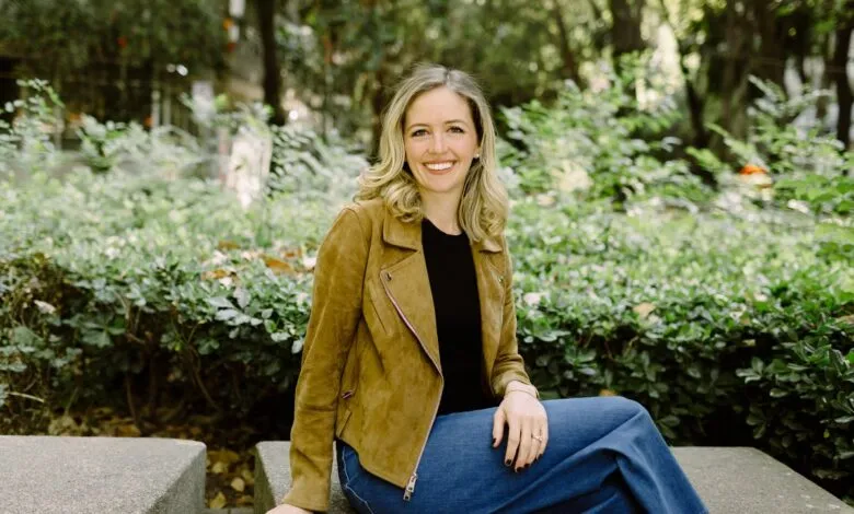 Smiling woman with blonde hair sits outdoors in a brown jacket and jeans.