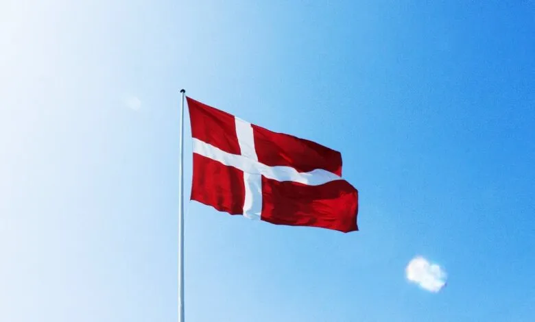 The Danish flag, a white Nordic cross on red, waving against a clear blue sky.