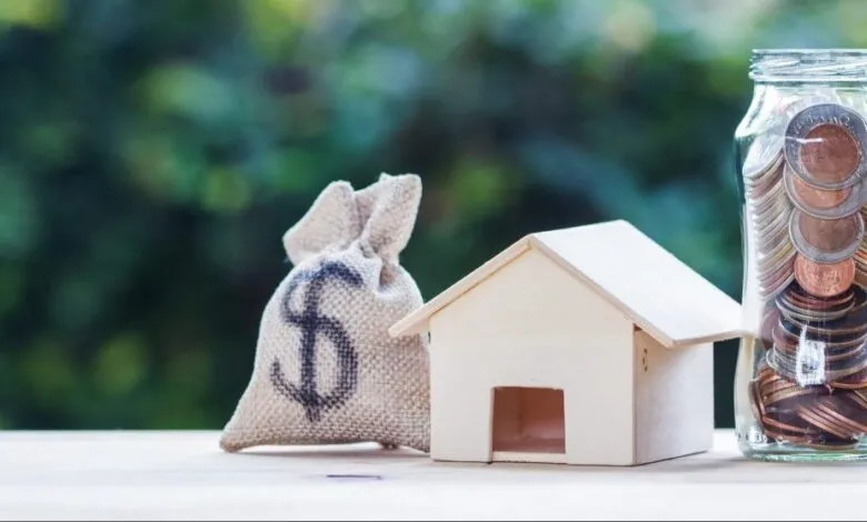 Money bag, toy house, and jar of coins on a wooden surface.