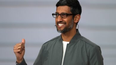 Sundar Pichai smiles, giving a thumbs up, wearing glasses and a gray jacket.
