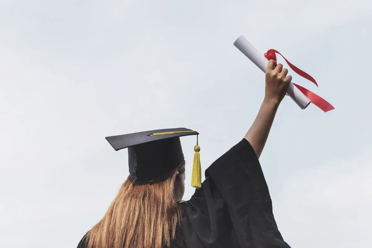 Graduate in cap and gown holds diploma high against sky.