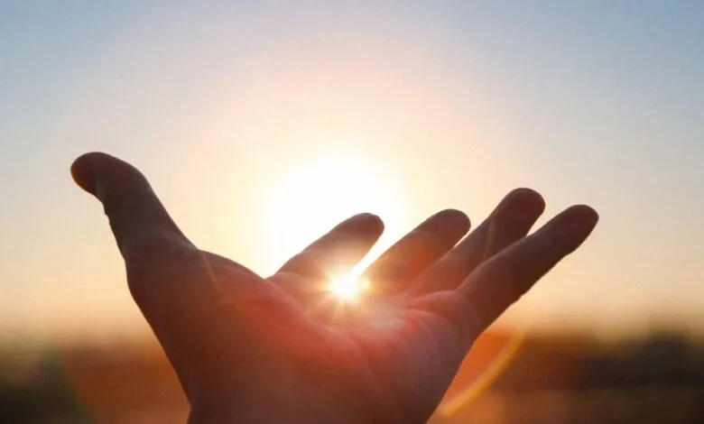 Hand reaching towards the sun, fingers slightly spread, against a soft, gradient sky.