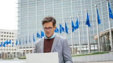 Man works on laptop with EU flags and building in background.