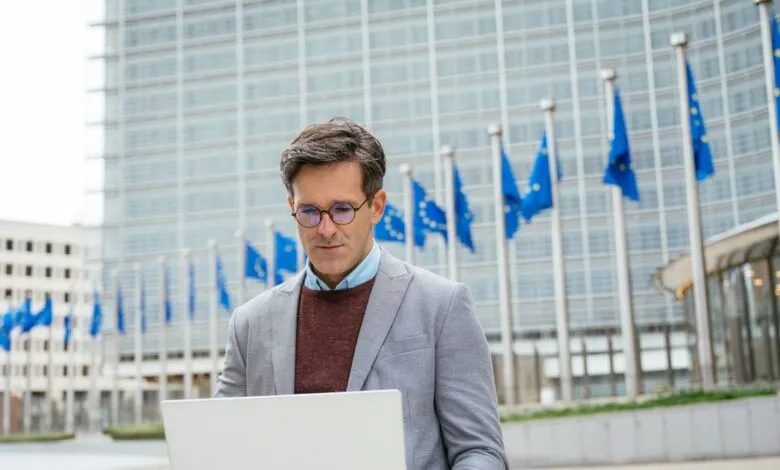 Man works on laptop with EU flags and building in background.