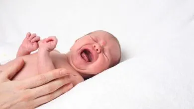 Crying baby lies on white blanket, held by a hand.