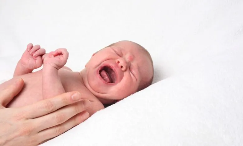 Crying baby lies on white blanket, held by a hand.