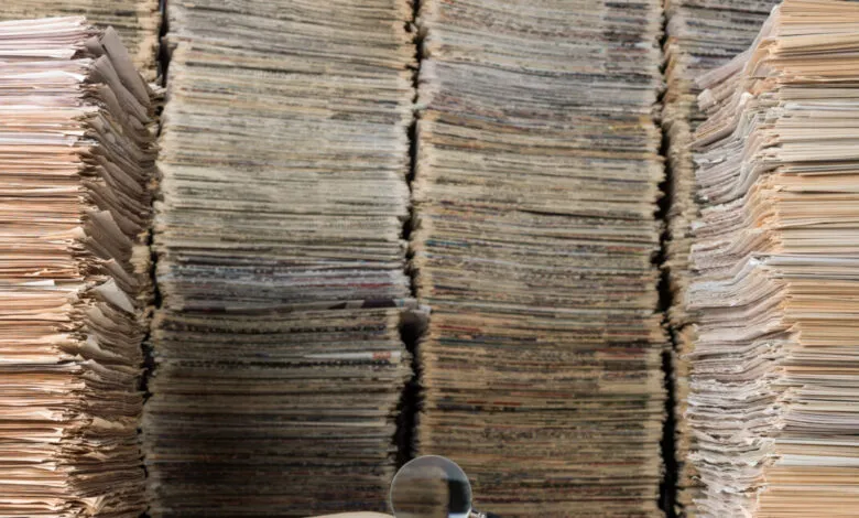 Stacks of aged documents, newspapers, and books with magnifying glass.
