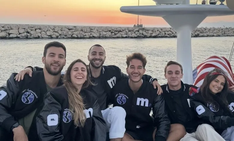 Group of six smiling people on a boat at sunset, wearing matching jackets.