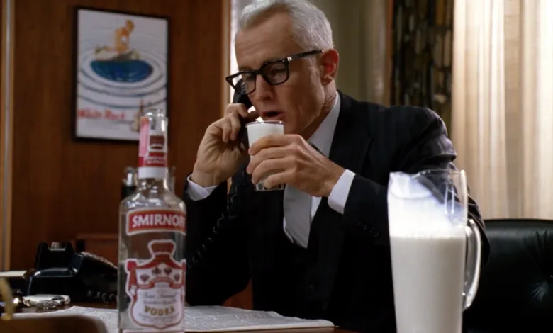 Man in suit drinking milk while on phone, Smirnoff bottle on desk.