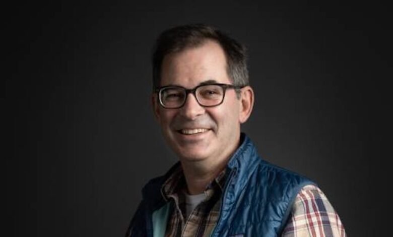 Headshot of a smiling man with glasses and a blue vest.