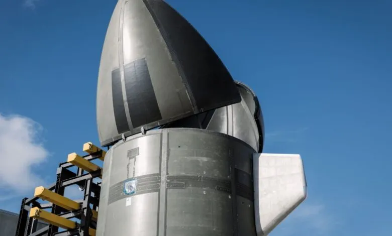 Close-up of a rocket nose cone with black heat shield tiles against a blue sky.