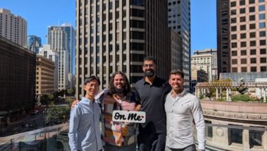 Four people pose with an 'on me' sign against a city skyline backdrop.