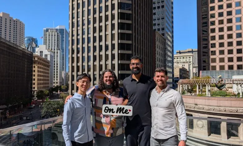 Four people pose with an 'on me' sign against a city skyline backdrop.