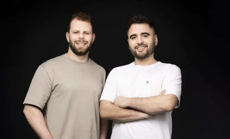 Two smiling men stand side-by-side against a dark background.