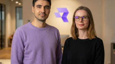 Two individuals stand in front of a logo, one in purple, the other in black.