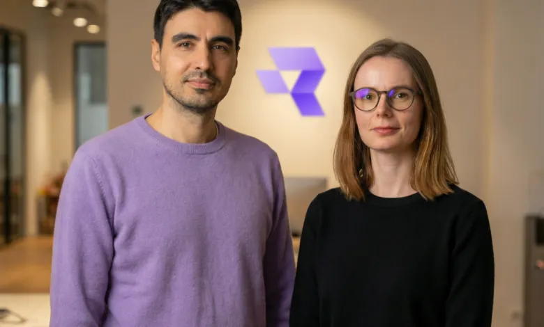 Two individuals stand in front of a logo, one in purple, the other in black.