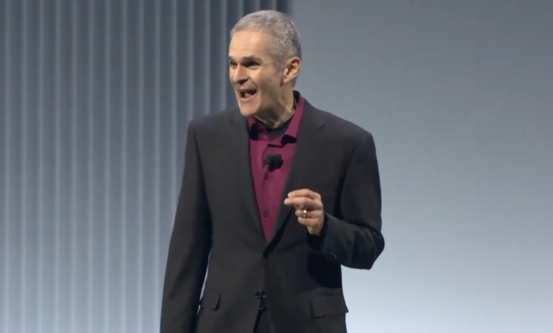 Man in suit gesturing while speaking on stage at a presentation.