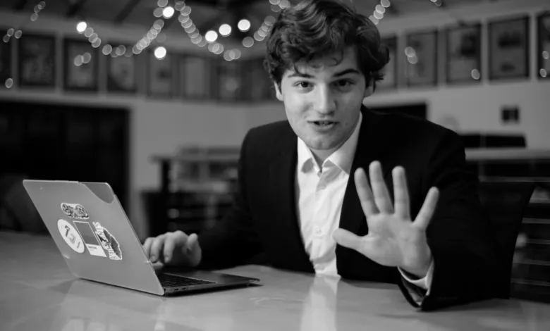 Young man in suit gestures while working on laptop at a table.