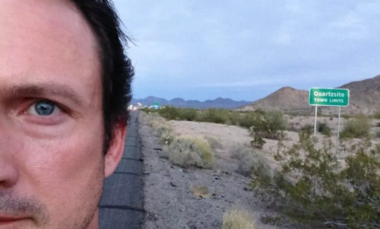 Man's selfie near Quartzsite town limits sign, desert landscape.