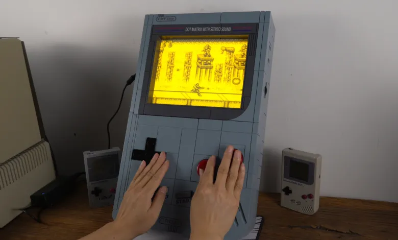 Oversized Game Boy replica with hands playing a game, smaller Game Boys nearby.