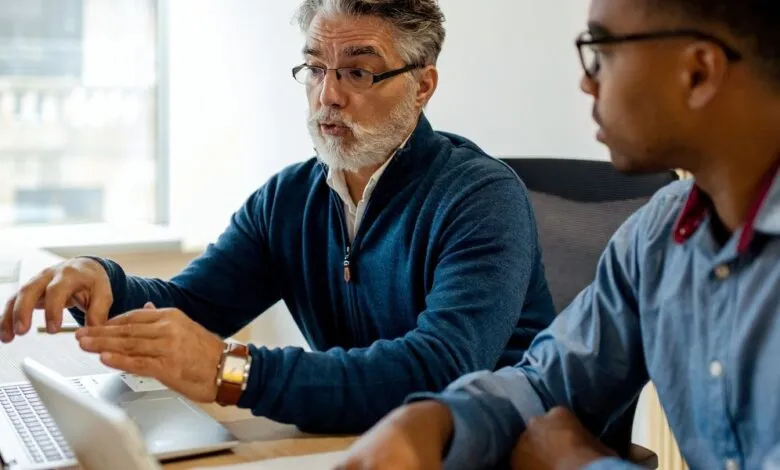 Two colleagues, one older with gray beard, working together at a desk.