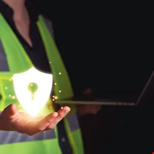 Person in safety vest holding glowing shield icon with keyhole, laptop in background.