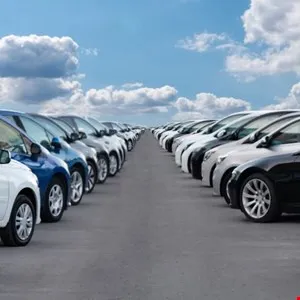 Rows of new cars parked on asphalt under a bright blue sky with fluffy white clouds.