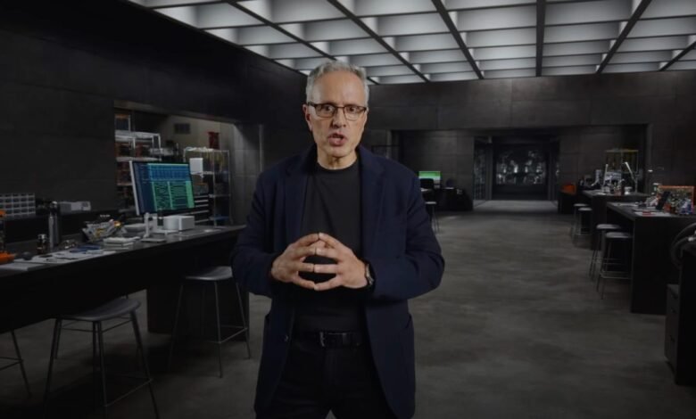Man with glasses stands in a high-tech lab, hands clasped, speaking.