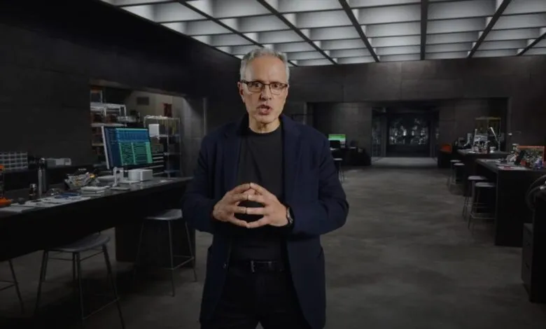 Man with glasses stands in a high-tech lab, hands clasped, speaking.