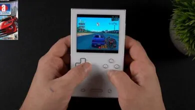 Hands play racing game on handheld console, 'Auto Modellista' box visible.