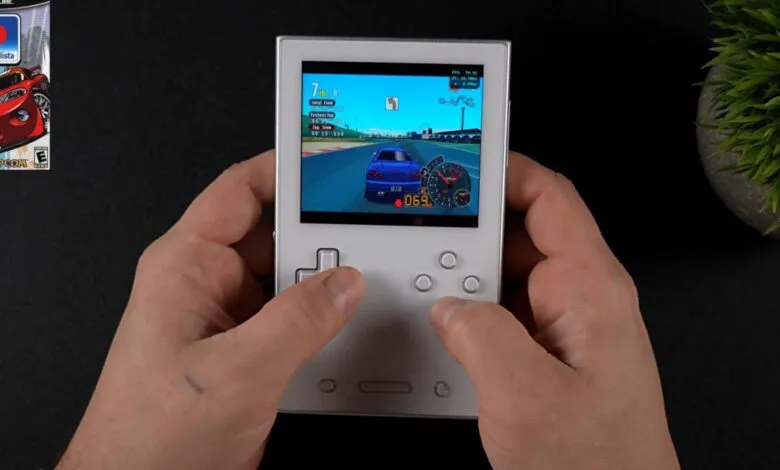Hands play racing game on handheld console, 'Auto Modellista' box visible.
