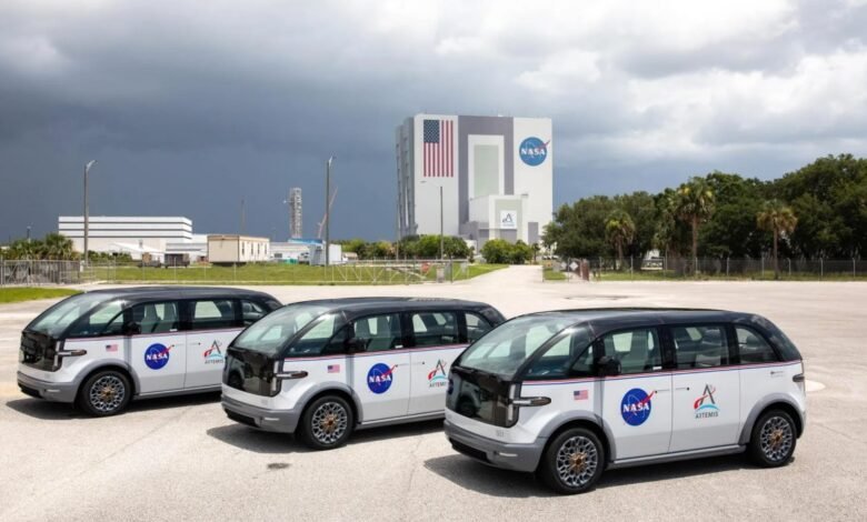 Three Canoo vehicles with NASA and Artemis logos parked in front of the Vehicle Assembly Building.