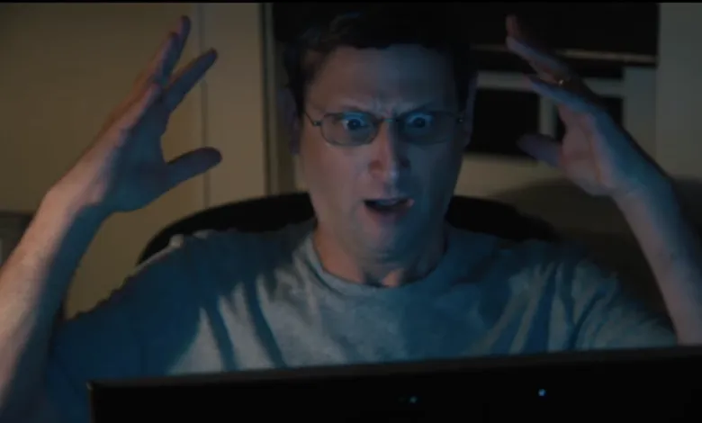 Man with glasses stares intensely at computer screen, hands raised in frustration.