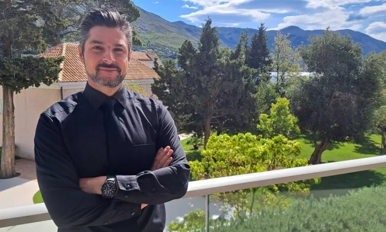 Man in black shirt stands on balcony with arms crossed, scenic backdrop.