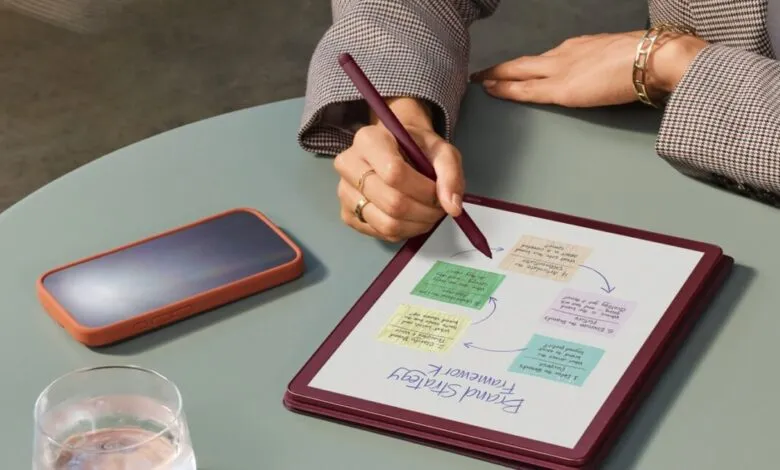 Person writing on tablet with stylus, phone and water glass on table.