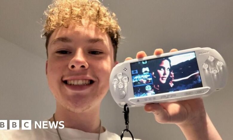 Smiling young man holds up a white PSP showing a video game.