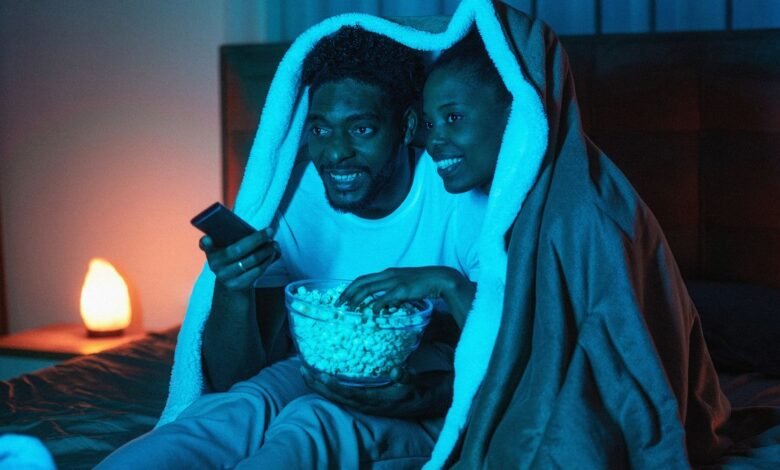 Couple under blanket enjoys movie night with popcorn and a phone.