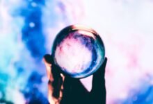 Hand holding crystal ball with galaxy inside against colorful background.