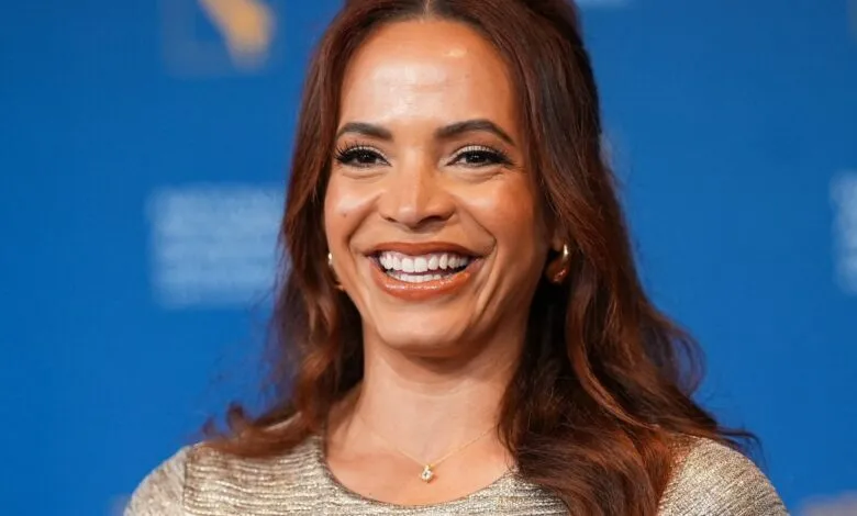 Sage Steele smiles against a blue backdrop, wearing gold jewelry and a shimmering top.