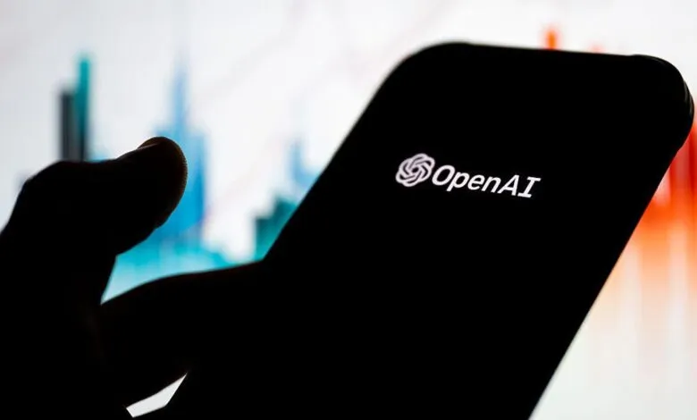 Close-up of a phone displaying the OpenAI logo, held by a silhouetted hand.