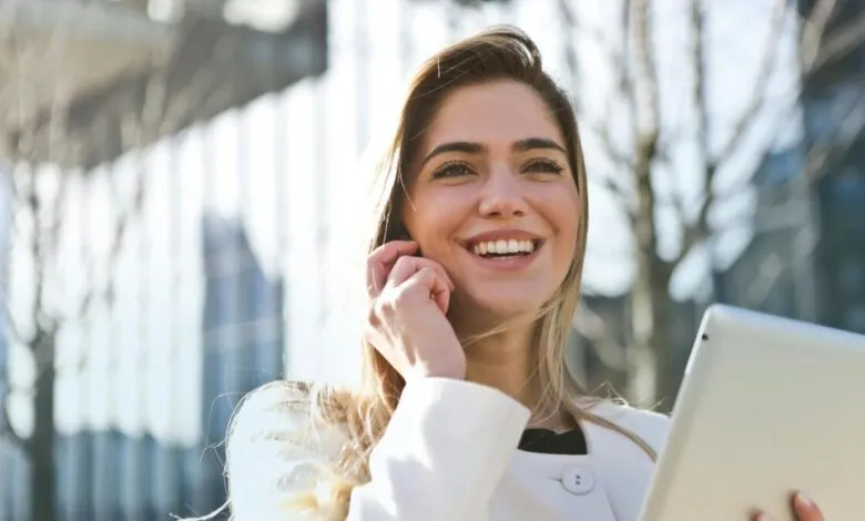 Smiling woman in white coat holds tablet outdoors.