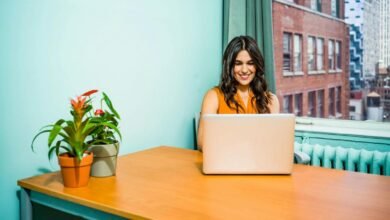 Woman smiling while working on laptop at desk with city view.