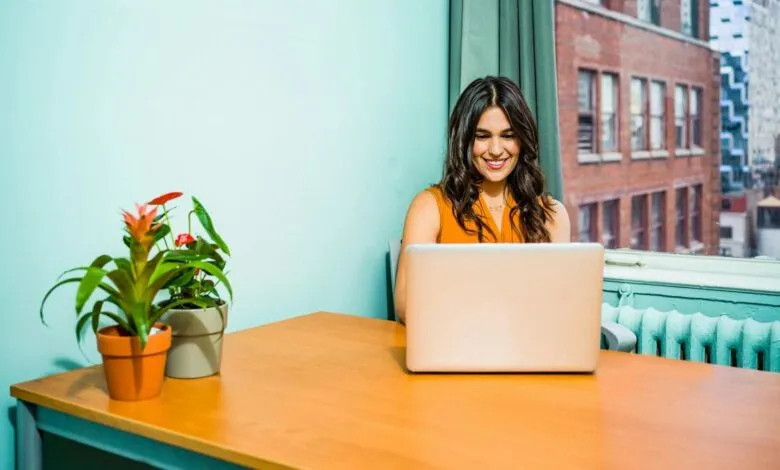 Woman smiling while working on laptop at desk with city view.