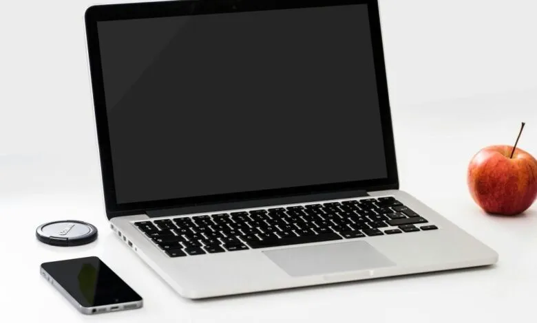 Laptop with black screen, phone, lens cap, and apple on white surface.