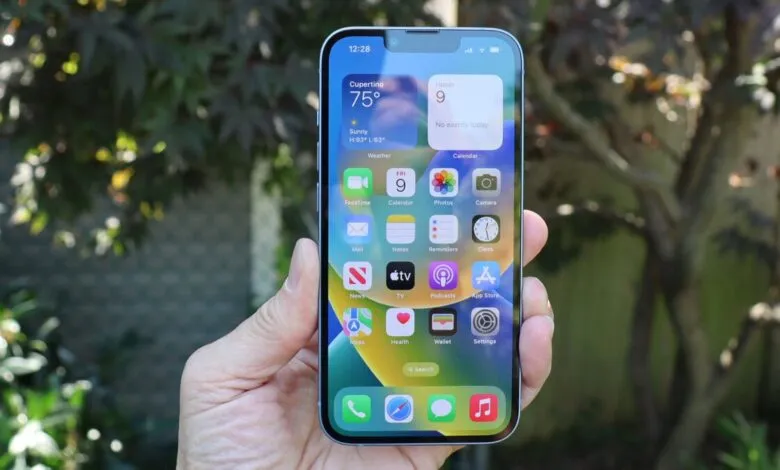 Hand holding iPhone displaying iOS 16 home screen with weather and app icons.