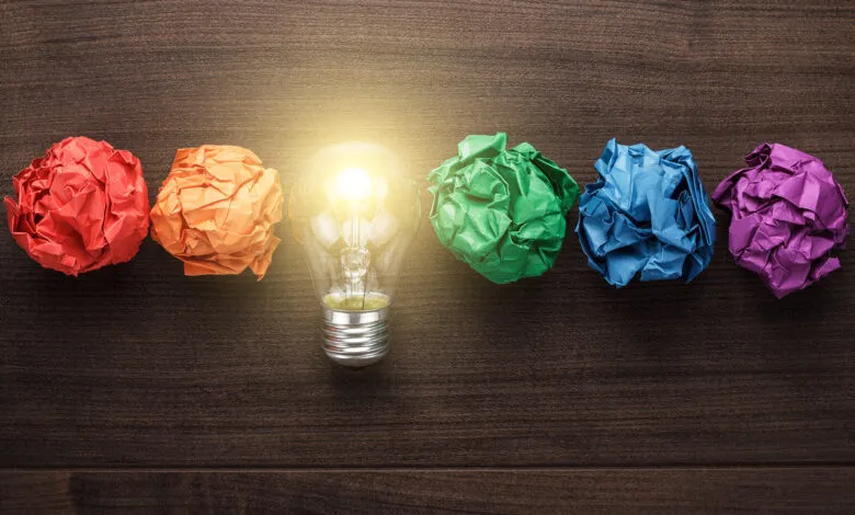 Bright lightbulb among colorful crumpled paper balls on wood.