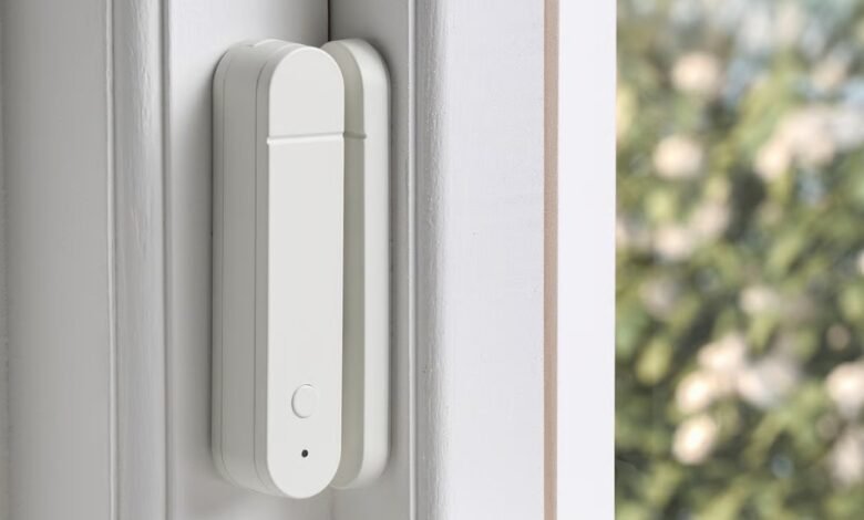 White smart sensor attached to a white window frame, blurred greenery visible outside.