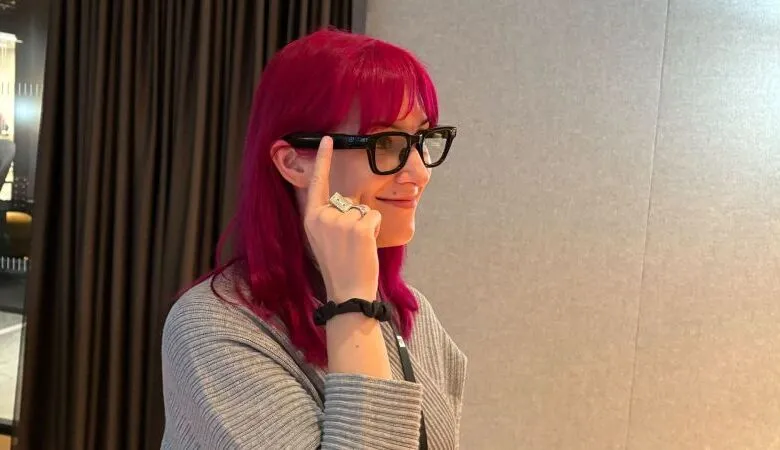 Woman with pink hair wearing smart glasses and pointing to the side.