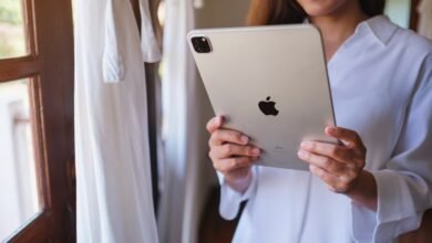 Woman holds a silver iPad near a window with white curtains.